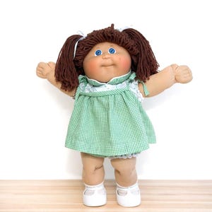 May include: A vintage Cabbage Patch Kid doll with brown yarn hair styled in pigtails, blue eyes, and rosy cheeks. The doll wears a green and white checkered dress with a white lace collar and matching white shoes. The doll's arms are outstretched.
