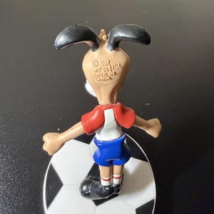 May include: A vintage figurine of a dog wearing a soccer uniform, standing on a soccer ball. The dog has black ears, a tan face, a red and white shirt, and blue shorts. The figurine is marked "Made in China" and "&copy; 1992".