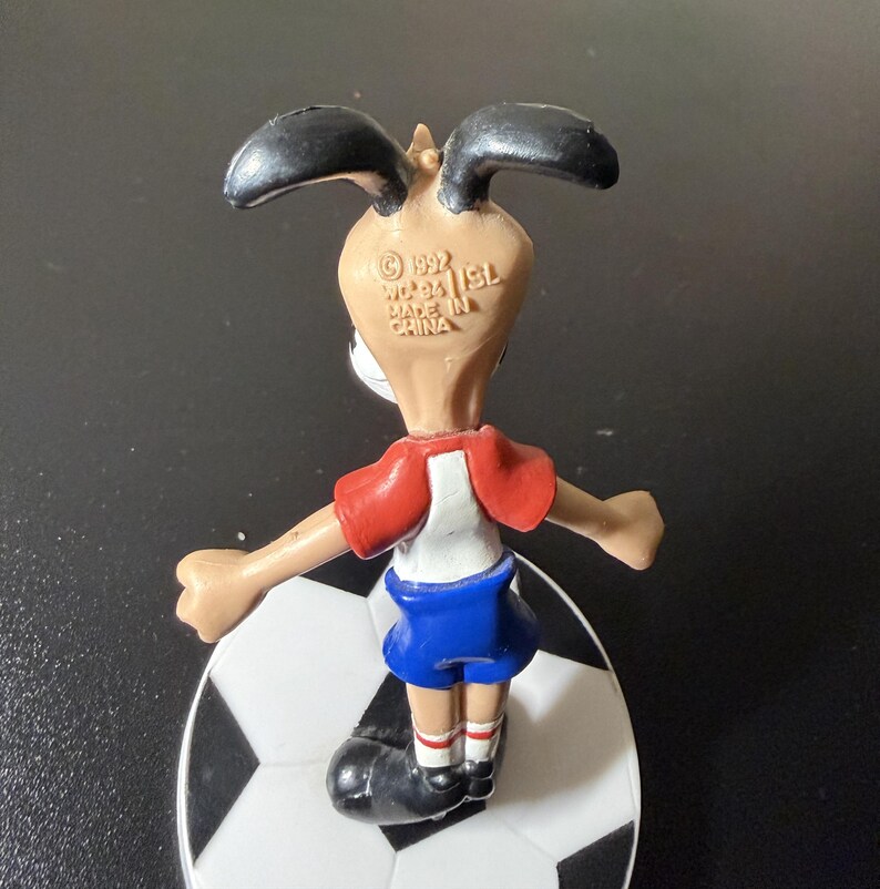 May include: A vintage figurine of a dog wearing a soccer uniform, standing on a soccer ball. The dog has black ears, a tan face, a red and white shirt, and blue shorts. The figurine is marked "Made in China" and "&copy; 1992".