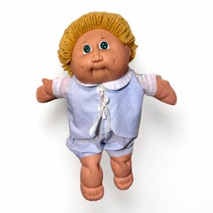 May include: A vintage Cabbage Patch Kid doll with a soft, vinyl face, green eyes, and a tuft of yellow yarn hair. The doll is wearing a blue and white checkered outfit with a white collar and matching shorts. The doll's arms are outstretched.