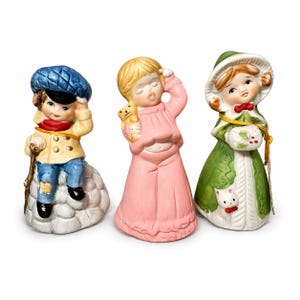 May include: Three ceramic figurines: a boy in a blue cap and yellow coat, a girl in a pink nightgown holding a teddy bear, and a girl in a green coat and hat. The figurines have painted details and are on a white background.