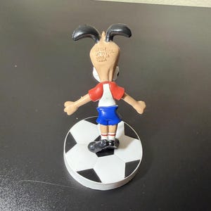 May include: A small figurine of a cartoon dog wearing a red and white shirt and blue shorts, standing on a soccer ball. The dog has black ears and is facing away from the viewer. The soccer ball is black and white.
