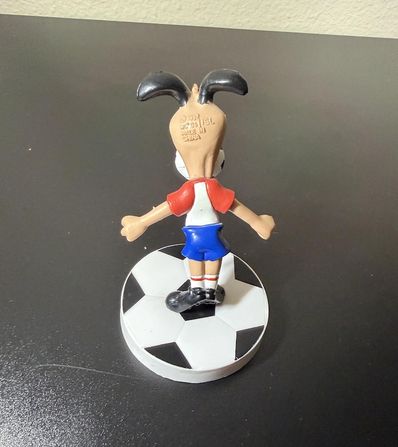 May include: A small figurine of a cartoon dog wearing a red and white shirt and blue shorts, standing on a soccer ball. The dog has black ears and is facing away from the viewer. The soccer ball is black and white.
