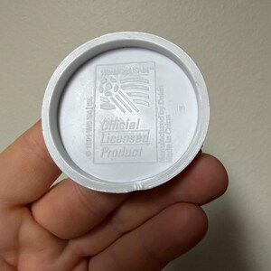May include: A white plastic disc with the text "World Cup USA94" and "Official Licensed Product." The disc has a circular design with a raised edge and is held in a hand.
