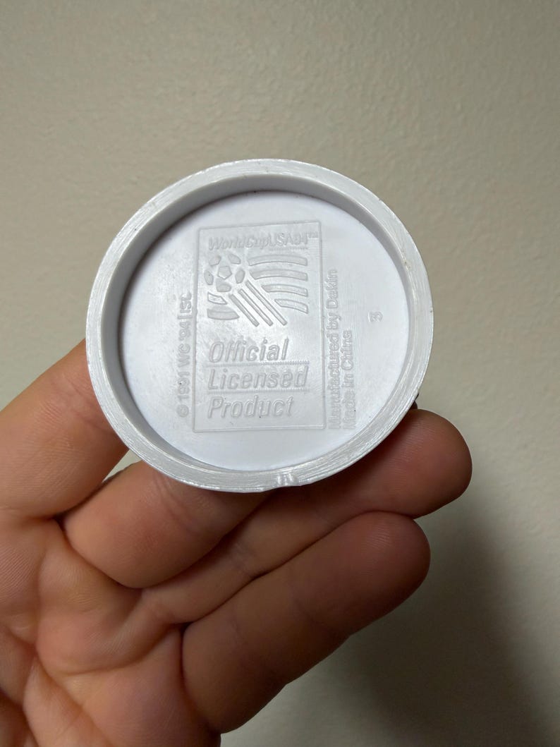May include: A white plastic disc with the text "World Cup USA94" and "Official Licensed Product." The disc has a circular design with a raised edge and is held in a hand.