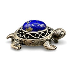 Può includere: Una spilla a forma di tartaruga color argento con un cabochon ovale di lapislazzuli lucido incastonato nel guscio. Il guscio in metallo presenta intricati motivi a volute, mentre la testa e le zampe sono dettagliate con piccoli punti in rilievo.