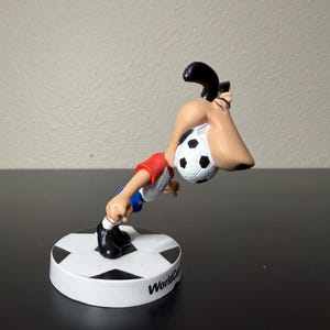 May include: A figurine of a cartoon character playing soccer. The character is wearing a red and white shirt, blue shorts, and black shoes. The soccer ball is black and white. The base is white with a black and white soccer ball design and the words "World Cup".