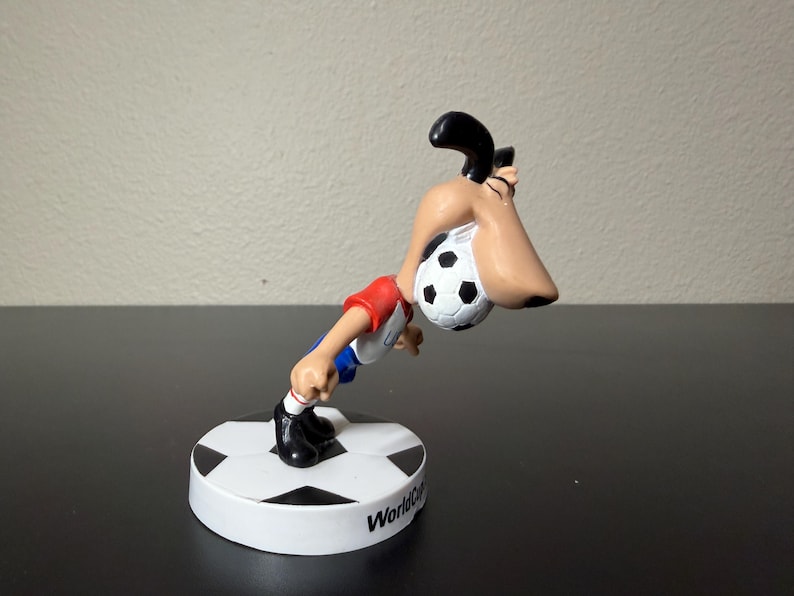 May include: A figurine of a cartoon character playing soccer. The character is wearing a red and white shirt, blue shorts, and black shoes. The soccer ball is black and white. The base is white with a black and white soccer ball design and the words "World Cup".