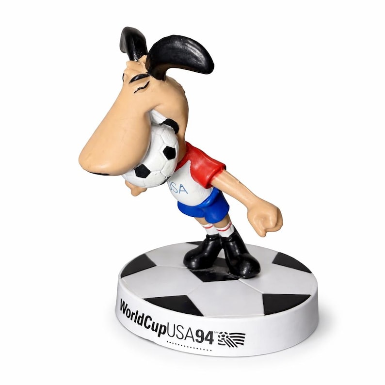 May include: A figurine of a cartoon dog mascot for the 1994 FIFA World Cup USA. The dog is wearing a red and blue uniform and holding a soccer ball. The base is a soccer ball with the text "WorldCupUSA94".