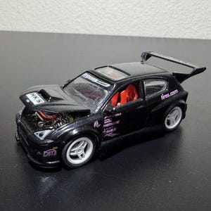 Könnte beinhalten: Ein schwarzes Spielzeugauto mit großem Heckflügel und weißen Rädern. Die Motorhaube ist geöffnet und gibt den Motor frei. Das Auto trägt den Text "tires.com" und "HKS". Der Innenraum ist rot.