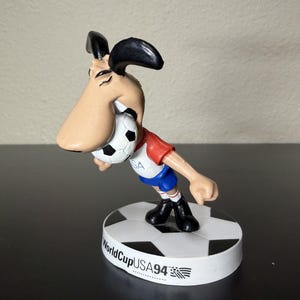 May include: A figurine of a cartoon dog in a red and blue soccer uniform, biting a soccer ball. The figurine is standing on a base with the text "World Cup USA 94".