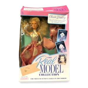 Puede incluir: Muñeca "The Real Model Collection" en caja, con Christie Brinkley. La muñeca lleva un top plateado y una falda rosa con lunares dorados. La caja incluye un bolso rosa, un folleto y una foto autografiada.