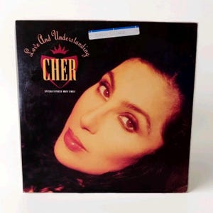 May include: Album cover for Cher's single, "Love and Understanding." The cover features a close-up of Cher's face with dark hair and red lipstick. The album title and artist name are in gold lettering.