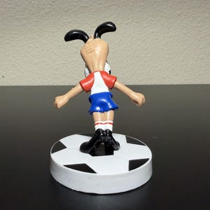 May include: A figurine of a cartoon dog in a soccer uniform, standing on a soccer ball-shaped base. The dog wears a red and white shirt, blue shorts, and black shoes. The base is white with black pentagons.