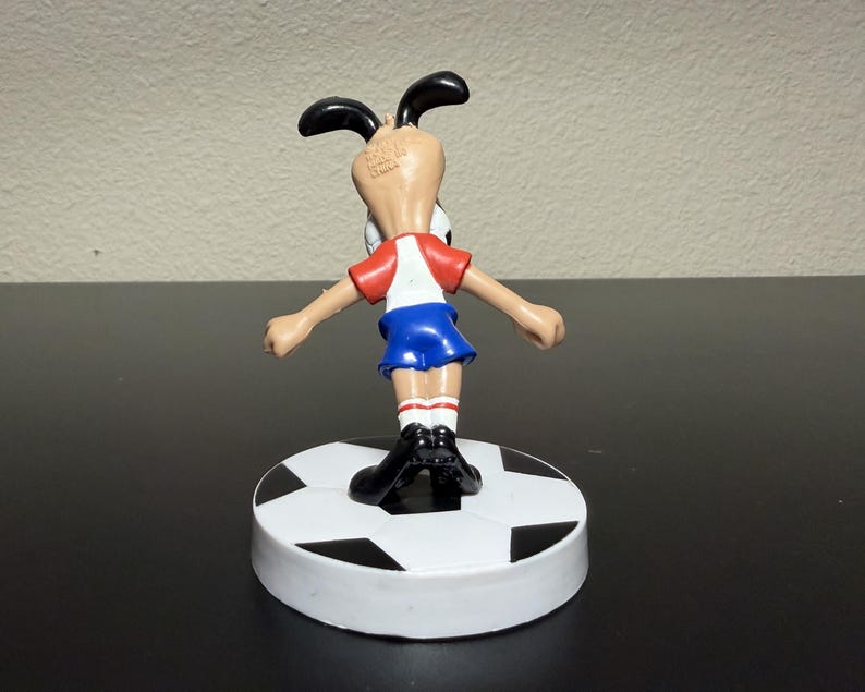 May include: A figurine of a cartoon dog in a soccer uniform, standing on a soccer ball-shaped base. The dog wears a red and white shirt, blue shorts, and black shoes. The base is white with black pentagons.