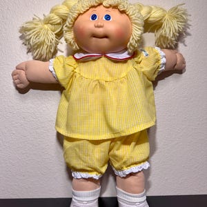 May include: A vintage doll with a soft, fabric body and a vinyl head. The doll has blonde yarn pigtails, blue eyes, and rosy cheeks. It wears a yellow gingham dress and shorts with white lace trim, white socks, and white shoes.