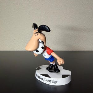 May include: A figurine of a cartoon dog wearing a red and blue soccer uniform, holding a soccer ball in its mouth. The figurine is standing on a base that says "WorldCupUSA94". The dog has a large nose and black ears.