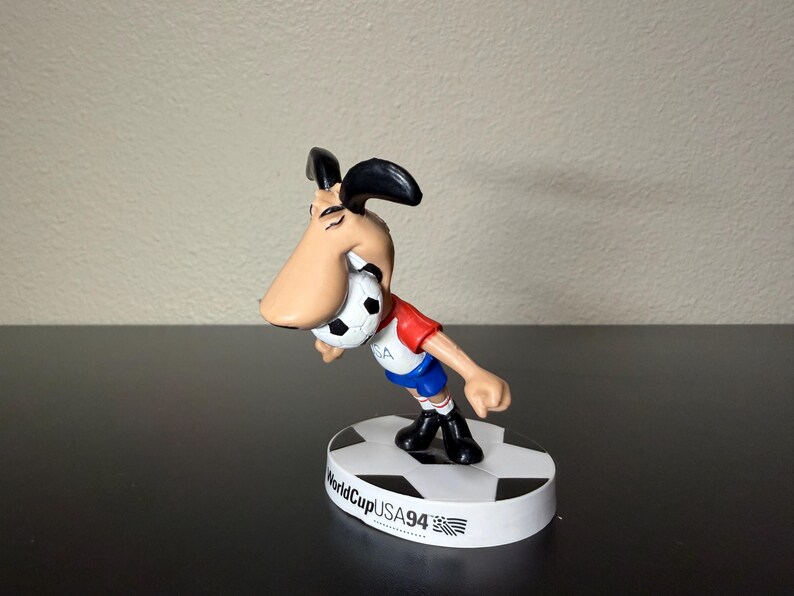 May include: A figurine of a cartoon dog wearing a red and blue soccer uniform, holding a soccer ball in its mouth. The figurine is standing on a base that says "WorldCupUSA94". The dog has a large nose and black ears.