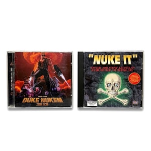 Op de afbeelding: Twee cd-rom-hoesjes naast elkaar. De linkerhoes toont een gespierde man met een geweer, de tekst "DUKE NUKEM 3D" en "USER GUIDE". De rechterhoes heeft een schedel en gekruiste botten met de tekst "NUKE IT".