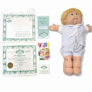 Puede incluir: Una muñeca Cabbage Patch Kids con cabello rubio de hilo, ojos verdes y un atuendo a cuadros azul y blanco. La muñeca está acompañada de papeles de adopción, un certificado de nacimiento y otros documentos relacionados.