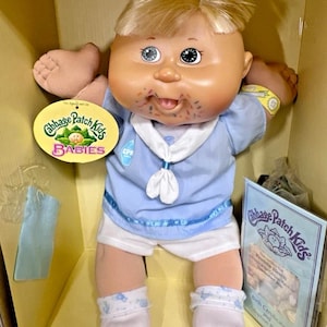 Könnte beinhalten: Cabbage Patch Kids Babies Puppe in einer Box. Die Puppe hat blondes Haar, blaue Augen und ein hellblaues Outfit im Matrosenstil mit weißen Shorts und Socken. Die Puppe hat ein Etikett mit der Aufschrift "Cabbage Patch Kids BABIES".
