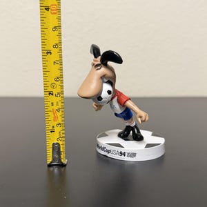 May include: A figurine of the 1994 World Cup USA mascot, a dog-like character, is shown. The figure is wearing a red and white shirt, blue shorts, and black shoes. The figurine is approximately 3 inches tall and stands on a white base.