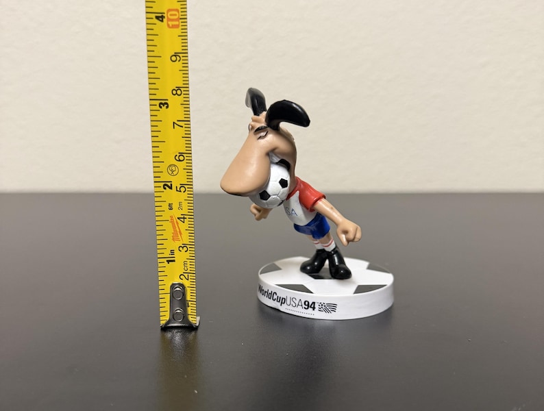 May include: A figurine of the 1994 World Cup USA mascot, a dog-like character, is shown. The figure is wearing a red and white shirt, blue shorts, and black shoes. The figurine is approximately 3 inches tall and stands on a white base.