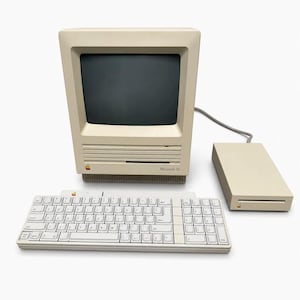 Peut inclure: Un ordinateur Macintosh SE beige avec un écran intégré, un lecteur de disquette et un clavier. L'ordinateur présente un petit logo Apple coloré et le texte Macintosh SE sur la façade. Un lecteur de disquette externe est relié par un câble.