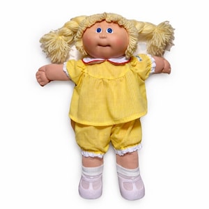 Puede incluir: Una muñeca vintage con coletas rubias de hilo, ojos azules y mejillas rosadas. La muñeca viste un conjunto de cuadros amarillos con ribete de encaje blanco, un cuello blanco con ribete rojo, calcetines blancos y zapatos lavanda. Un juguete clásico.