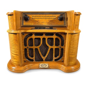 May include: A wooden tabletop radio featuring a light brown finish with a prominent speaker grille in the centre. The upper section includes a horizontal dial with AM and FM frequency markings, two black control knobs, and a world map background.