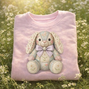 Peut inclure: Un sweat-shirt rose clair avec un appliqué de lapin décoratif. Le lapin est bleu clair avec des motifs floraux, des oreilles bordées de dentelle et un nœud lavande. Les pieds du lapin ont des motifs de marguerites. Le sweat-shirt est plié sur un lit de fleurs blanches.