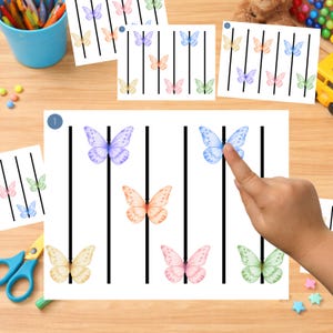 May include: White paper with colorful watercolor butterflies and black vertical lines. A finger points to a blue butterfly. Other papers with butterflies and lines are scattered around. Scissors and a blue cup are also visible.
