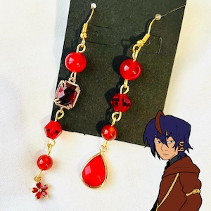以下が含まれることがあります： 赤いビーズと宝石が付いた、ぶら下がりタイプのイヤリングのペア。イヤリングはゴールドカラーのフックとアクセントが付いています。片方のイヤリングには涙型の宝石、もう片方には四角いカットの宝石が付いています。小さな赤い花のチャームが片方のイヤリングの下部にあります。