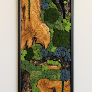 Op de afbeelding: Een ingelijst wandkunstwerk met een natuurlijk ontwerp. Het toont houtnerf, groene en blauwe mossen en een verticaal groen, getextureerd element. Het kunstwerk is gevat in een zwarte lijst. Afmetingen: ongeveer 30 cm breed en 60 cm hoog.