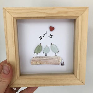 May include: A square light wood frame containing a white background with three birds made of sea glass perched on a piece of driftwood. Above the birds are black musical notes and a small red heart. The name Nikki is signed in black ink.