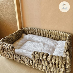 May include: A rectangular pet bed with a woven, light brown exterior and a fluffy, white interior. The bed has a low profile with a raised edge and is designed for small pets. The bed is made of natural materials.