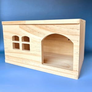 May include: A natural wood pet house with an arched doorway and two small square windows. The house is rectangular and appears to be made of unfinished wood. The background is a solid blue.