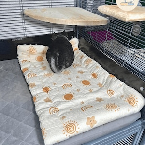 May include: A small pet, possibly a chinchilla, rests on a patterned pet bed inside a wire cage. The bed is white with a repeating pattern of suns, rainbows, and stars in a warm brown color. Wooden shelves are mounted on the cage walls.