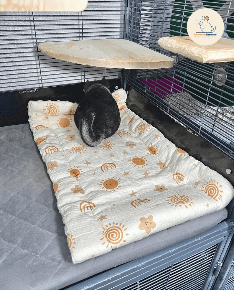 May include: A small pet, possibly a chinchilla, rests on a patterned pet bed inside a wire cage. The bed is white with a repeating pattern of suns, rainbows, and stars in a warm brown color. Wooden shelves are mounted on the cage walls.