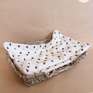 Puede incluir: Una cesta rectangular de mimbre trenzado que contiene un forro de tela color crema suave con un patr&oacute;n repetitivo de peque&ntilde;os corazones marrones. El forro presenta una textura acolchada y est&aacute; ajustado al interior de la cesta de fibra natural.