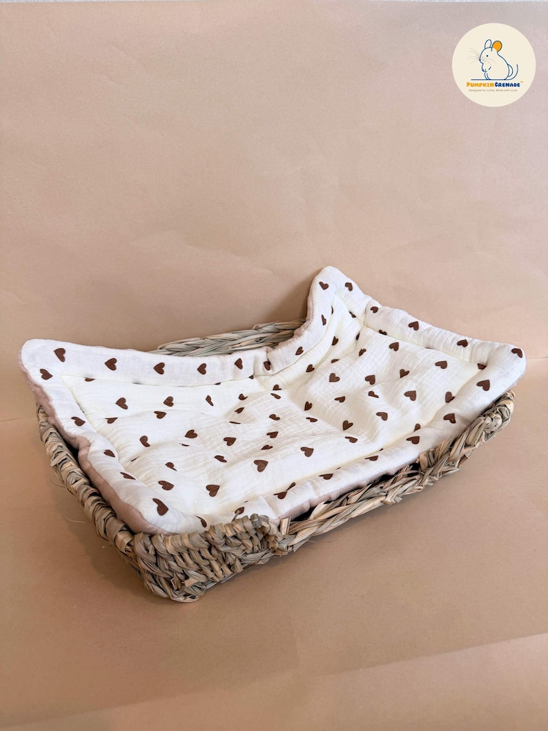 Puede incluir: Una cesta rectangular de mimbre trenzado que contiene un forro de tela color crema suave con un patr&oacute;n repetitivo de peque&ntilde;os corazones marrones. El forro presenta una textura acolchada y est&aacute; ajustado al interior de la cesta de fibra natural.