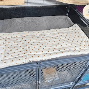 May include: A rectangular pet bed with a cream-colored surface adorned with small brown hearts. The bed is quilted and sits inside a metal cage, providing a comfortable space for small animals. The cage has a gray padded area.