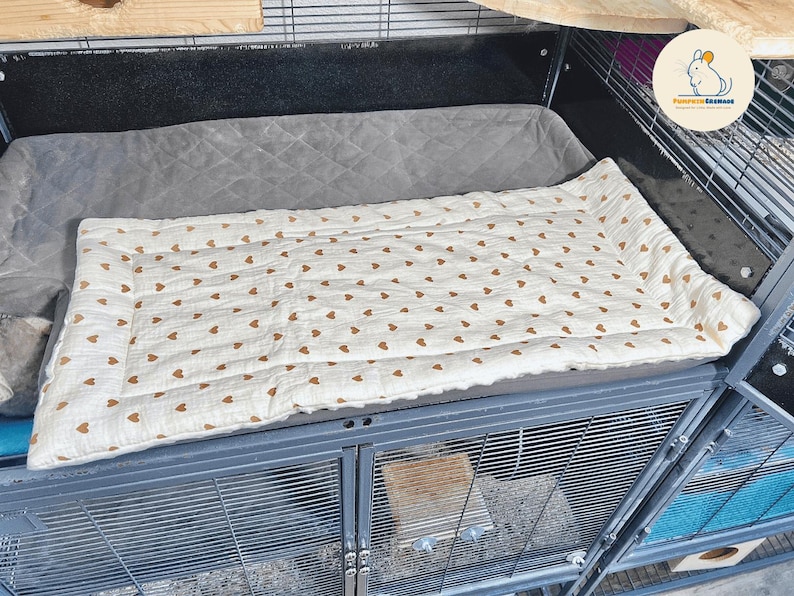 May include: A rectangular pet bed with a cream-colored surface adorned with small brown hearts. The bed is quilted and sits inside a metal cage, providing a comfortable space for small animals. The cage has a gray padded area.
