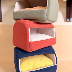 Puede incluir: Una pila de tres camas para mascotas cubiertas de forro polar suave con frentes abiertos. La cama superior es verde salvia con cojín naranja, la del medio es roja con cojín blanco y la inferior es azul marino con cojín amarillo. Logo PUMPKIN CREMAGE.