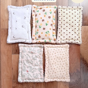 Puede incluir: Cinco cojines rectangulares de algod&oacute;n con esquinas redondeadas y bordes acolchados. Los patrones incluyen blanco con lunas oscuras, crema con osos marrones y tulipanes amarillos, crema con corazones marrones, follaje verde y peque&ntilde;os estampados florales.