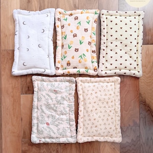 Puede incluir: Cinco cojines rectangulares de muselina con esquinas redondeadas y bordes cosidos. Los estampados incluyen blanco con lunas grises, crema con osos marrones y tulipanes amarillos, crema con corazones marrones, crema con follaje verde y flores peque&ntilde;as.