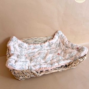Puede incluir: Una cesta rectangular de mimbre trenzado que contiene un forro de tela acolchada suave. El forro es de color blanco roto con un patr&oacute;n repetitivo de hojas verde salvia y peque&ntilde;as bayas rojas, con un borde de tela rosa claro s&oacute;lido.