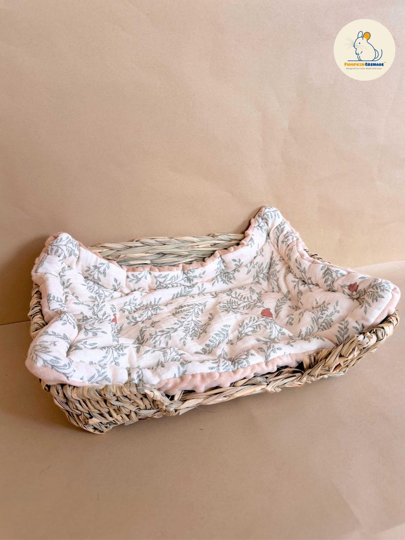 Puede incluir: Una cesta rectangular de mimbre trenzado que contiene un forro de tela acolchada suave. El forro es de color blanco roto con un patr&oacute;n repetitivo de hojas verde salvia y peque&ntilde;as bayas rojas, con un borde de tela rosa claro s&oacute;lido.