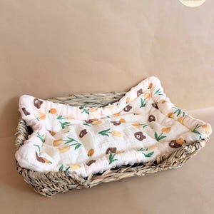 Puede incluir: Una cesta rectangular de pasto marino trenzado que contiene un forro de tela acolchada de color crema. El forro presenta un patr&oacute;n repetitivo de peque&ntilde;as caras de oso marr&oacute;n, flores amarillas tipo tulip&aacute;n y tallos verdes, con un borde acolchado.