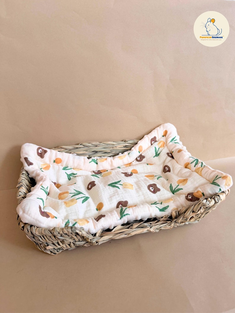 Puede incluir: Una cesta rectangular de pasto marino trenzado que contiene un forro de tela acolchada de color crema. El forro presenta un patr&oacute;n repetitivo de peque&ntilde;as caras de oso marr&oacute;n, flores amarillas tipo tulip&aacute;n y tallos verdes, con un borde acolchado.
